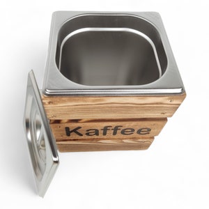 May include: A wooden box with a stainless steel container inside. The box has the word "Kaffee" printed on the front. The container has a lid that is open and sitting next to the box.