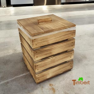 Könnte beinhalten: Ein quadratischer Mülleimer aus Holz mit Deckel und Plastikfutter. Der Mülleimer ist aus Naturholz mit rustikalem Finish. Der Mülleimer steht auf einem Betonboden.