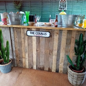 May include: A rustic wooden bar with a sign that reads "The Coxalls Est 18th August 2021". The bar is decorated with plants and a metal ice bucket.