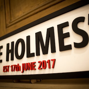May include: A white sign with black lettering that reads "THE HOLMES'S EST 17th JUNE 2017". The sign is made of wood and has a black frame.