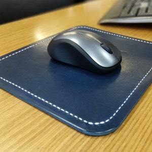 Könnte beinhalten: Ein dunkelblaues Leder-Mauspad mit weißen Nähten. Eine silberne kabellose Maus liegt auf dem Pad.