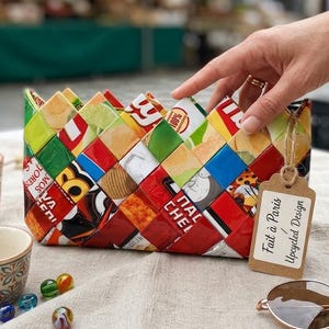 Peut inclure: Une pochette rectangulaire fabriquée à partir de bandes tressées d'emballages alimentaires recyclés colorés. Le sac présente un bord supérieur en zigzag et une étiquette en papier brun attachée avec de la ficelle portant le texte Fait à Paris / Upcycled Design.