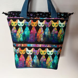 May include: A colorful tote bag with a repeating pattern of stylized cats in various colors. The cats are set against a black background with a denim blue handle. The top edge features a paw print design.