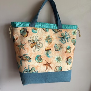 May include: A tote bag with a marine life pattern. The bag features a light peach background with illustrations of sea creatures in teal and blue. The top and bottom are teal and denim blue, with denim blue handles.