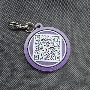 May include: A round, purple keychain tag with a white QR code in the center. The tag has a silver-colored clasp for attaching to keys or a bag. The QR code is a complex pattern of black and white squares.