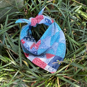 Può includere: Una bandana patchwork blu, rossa e bianca, annodata in alto. Il tessuto presenta vari motivi, tra cui disegni floreali e geometrici. La bandana è esposta su un letto di erba verde.