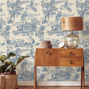 May include: A room featuring a cream-colored wallpaper with a blue toile pattern of animals and landscapes. A wooden side table with a lamp, clock, and decorative boxes sits next to the wallpaper. A woven basket with a plant is in the foreground.