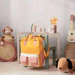 K&ouml;nnte beinhalten: Ein gelber Rucksack mit rosa Taschen h&auml;ngt an einem gr&uuml;nen Stuhl in einem Raum mit einer pink-wei&szlig;en Chevron-Tapete. Ein Holztisch mit einem Regenbogenspielzeug und einer sternf&ouml;rmigen Lampe steht vor dem Stuhl. Ein Stofftier-Affe sitzt in einem Weidenkorb auf dem Boden.