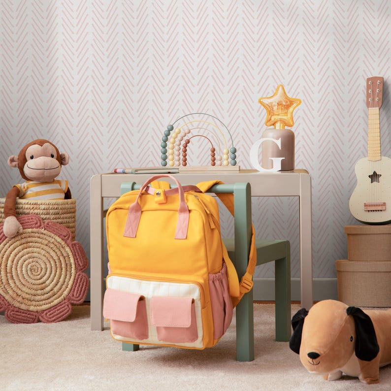 K&ouml;nnte beinhalten: Ein gelber Rucksack mit rosa Taschen h&auml;ngt an einem gr&uuml;nen Stuhl in einem Raum mit einer pink-wei&szlig;en Chevron-Tapete. Ein Holztisch mit einem Regenbogenspielzeug und einer sternf&ouml;rmigen Lampe steht vor dem Stuhl. Ein Stofftier-Affe sitzt in einem Weidenkorb auf dem Boden.