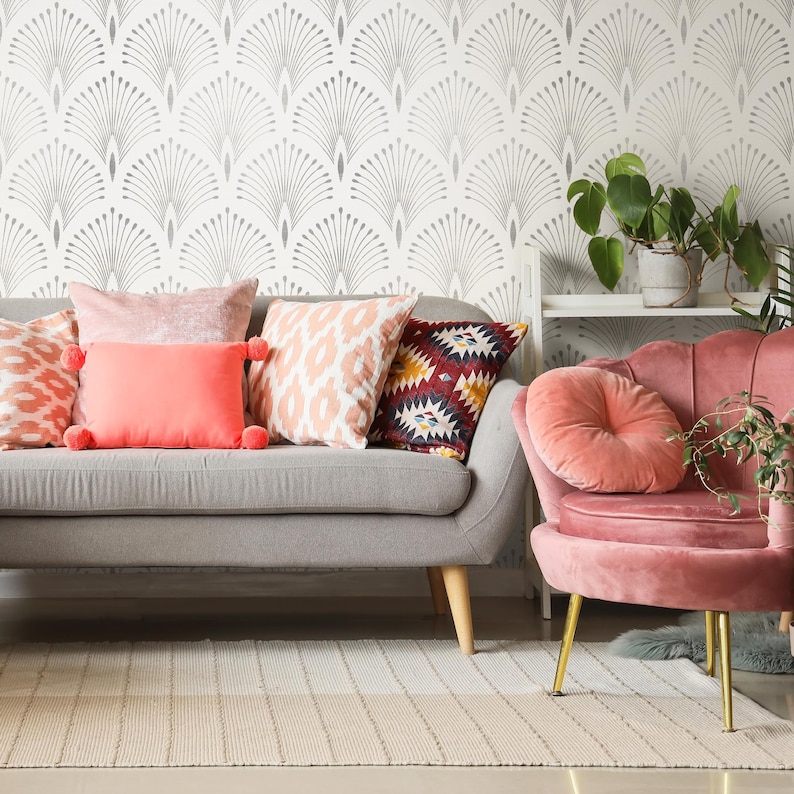 May include: A grey sofa with a variety of colourful throw pillows, including pink, red and orange. The sofa is in front of a white wall with a grey geometric pattern. A pink velvet armchair with a round pink cushion sits to the right of the sofa.