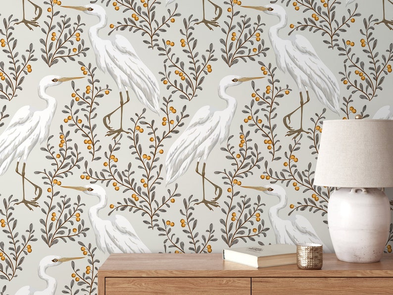 May include: A white background with a repeating pattern of white herons and grey branches with yellow berries. The pattern is on a wall behind a wooden table with a lamp and a book on it.