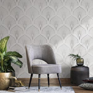 May include: A grey armchair with a white fabric seat and black legs sits in front of a white wall with a silver geometric pattern. A potted plant and a side table with a vase are also in the room.