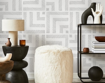 May include: A white faux fur ottoman with a black and white geometric patterned wallpaper in the background. A black metal shelf with a black vase and a bowl on it is to the right of the ottoman.