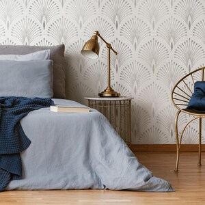 May include: A bedroom featuring a bed, a bedside table, a lamp, and a chair. The walls are adorned with a white and grey wallpaper featuring a geometric pattern. The bed has a blue blanket and a white pillow. The bedside table is made of metal and has a white marble top. The lamp is gold and has a white shade. The chair is made of metal and has a blue cushion.