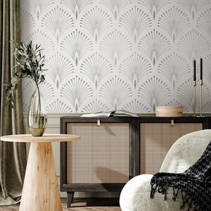 May include: A white wallpaper with a silver geometric pattern of stylised peacock feathers. The wallpaper is in a living room setting with a wooden side table, a chair, and a cabinet with woven cane doors.