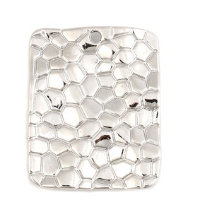 Puede incluir: Un colgante rectangular plateado con un patrón texturizado en forma de panal. El colgante tiene un pequeño agujero en la parte superior para una cadena o cordón. La superficie refleja la luz, creando reflejos y sombras.