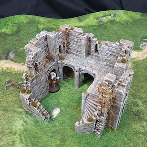 May include: A miniature, weathered stone castle ruin with arched doorways, staircases, and a small figure inside. The model is gray with patches of green and brown, set against a green and brown landscape.