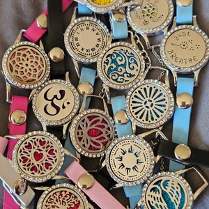 May include: A collection of 12 silver-toned aromatherapy diffuser bracelets with various designs and colors. The bracelets feature a variety of designs, including a butterfly, a clock, a flower, a tree, a heart, a sun, and wings. The bracelets are made of leather and have a silver-toned metal clasp.