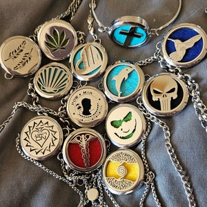 May include: A collection of silver-toned essential oil diffuser necklaces with various designs, including a seashell, a cross, a dolphin, a skull, a butterfly, a heart, a medical symbol, a tree, a cannabis leaf, and a hummingbird.  The necklaces are on a gray fabric background.