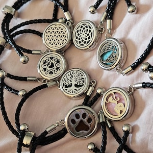 May include: Seven black braided bracelets with silver metal charms. Each charm has a different design, including a tree, a paw print, a heart, a cat, wings, and two geometric designs.