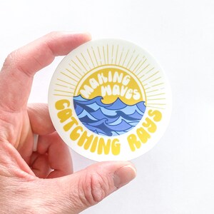 May include: A white round sticker with a yellow sunburst design. The sunburst has a yellow circle in the center with the text "Making Waves Catching Rays" in a retro font.