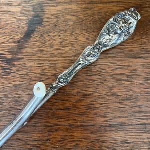 May include: An antique silver-plated serving utensil with an ornate handle featuring floral and face details. The utensil has a long, slender design with a small, circular attachment at the end. The piece is set against a wooden surface.