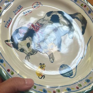 May include: A ceramic plate with a hand-painted illustration of a playful cat wearing a red bow. The plate has a scalloped edge with a floral and leaf pattern. Text in French reads "Viens jouer avec moi!"