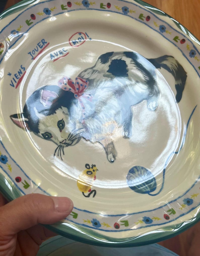 May include: A ceramic plate with a hand-painted illustration of a playful cat wearing a red bow. The plate has a scalloped edge with a floral and leaf pattern. Text in French reads "Viens jouer avec moi!"