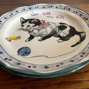 May include: A decorative ceramic plate with a hand-painted cat design. The cat is black and white with a pink bow, and the plate has a blue and green floral border. Text on the plate reads "Viens Jouer avec Moi!"