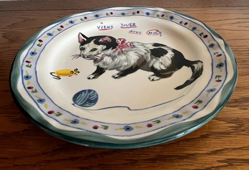 May include: A decorative ceramic plate with a hand-painted cat design. The cat is black and white with a pink bow, and the plate has a blue and green floral border. Text on the plate reads "Viens Jouer avec Moi!"