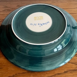 May include: A teal-colored ceramic plate with a white rim and a white center. The plate's underside features the artist's signature, "Nathalie Rete," and other text. The plate has a hand-painted appearance with visible brushstrokes.