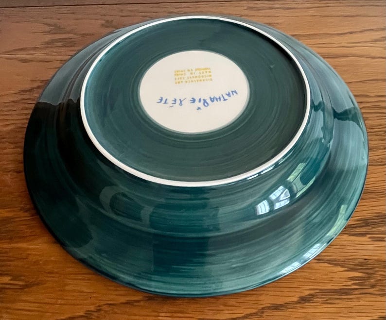 May include: A teal-colored ceramic plate with a white rim and a white center. The plate's underside features the artist's signature, "Nathalie Rete," and other text. The plate has a hand-painted appearance with visible brushstrokes.
