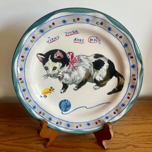 May include: A decorative ceramic plate featuring a hand-painted black and white cat with a red bow, a yellow mouse, and a blue ball of yarn. The plate has a blue and green floral border and the French text "Viens jouer avec moi!"