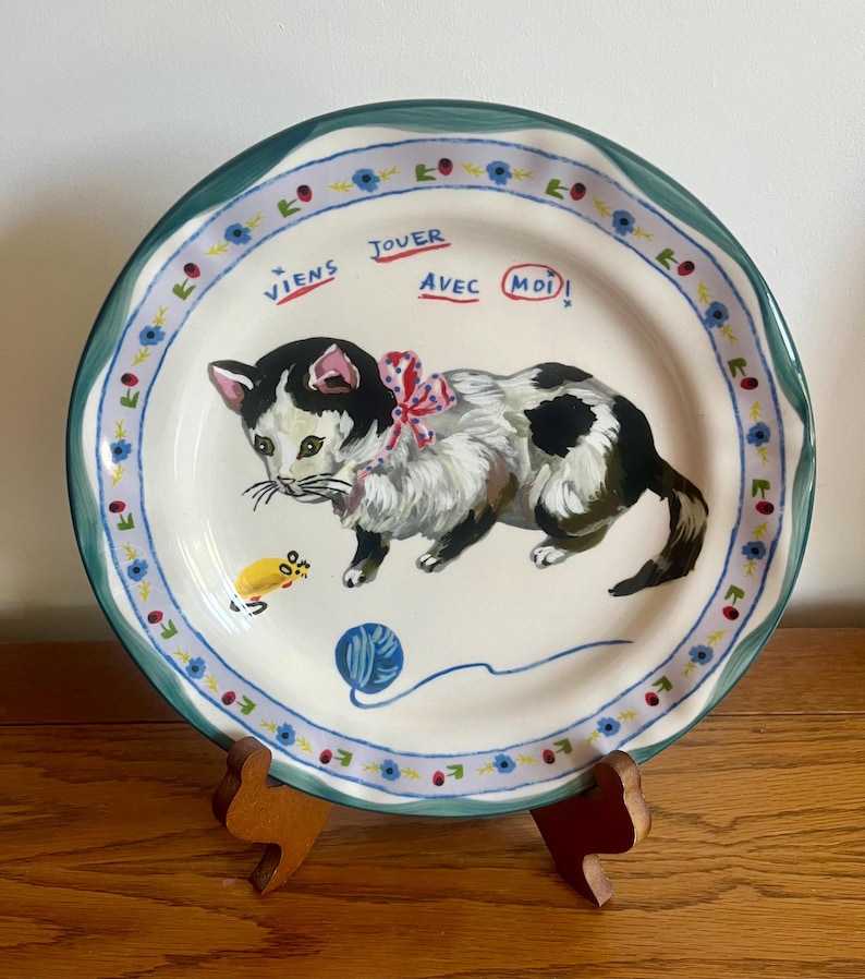 May include: A decorative ceramic plate featuring a hand-painted black and white cat with a red bow, a yellow mouse, and a blue ball of yarn. The plate has a blue and green floral border and the French text "Viens jouer avec moi!"