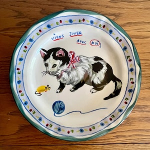 May include: A decorative ceramic plate featuring a playful cat design. The cat is black and white with a pink bow, a yellow mouse, and a blue yarn ball. The plate has a blue and green floral border and the text "Viens Jouer Avec Moi!"