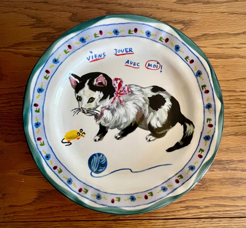 May include: A decorative ceramic plate featuring a playful cat design. The cat is black and white with a pink bow, a yellow mouse, and a blue yarn ball. The plate has a blue and green floral border and the text "Viens Jouer Avec Moi!"