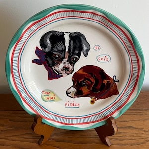 Może przedstawiać: Ozdobny ceramiczny talerz z dwoma portretami ps&oacute;w. Talerz ma białe tło z zieloną, czerwoną i niebieską obw&oacute;dką. Psy są namalowane w kolorach czarnym, białym i brązowym, z tekstem "Je Suis", "Ton Ami" i "Fidele".