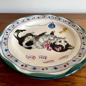 May include: A ceramic plate with a playful cat design. The cat is black and white, with a red bow, and is lying on its back. The plate has a green rim and a blue and white floral border. The words "Avec Moi! Viens Jouer" are written on the plate.