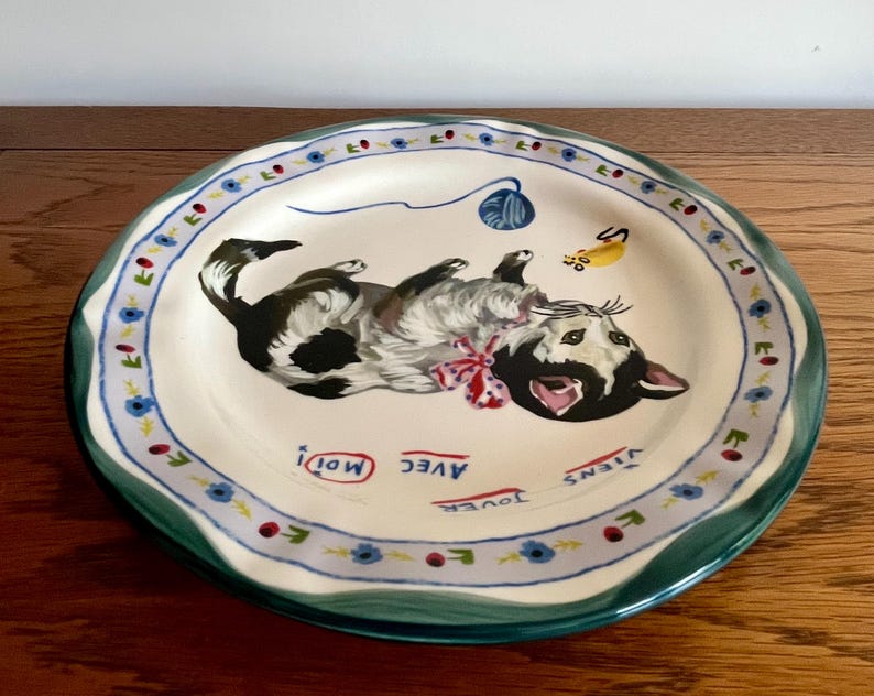 May include: A ceramic plate with a playful cat design. The cat is black and white, with a red bow, and is lying on its back. The plate has a green rim and a blue and white floral border. The words "Avec Moi! Viens Jouer" are written on the plate.