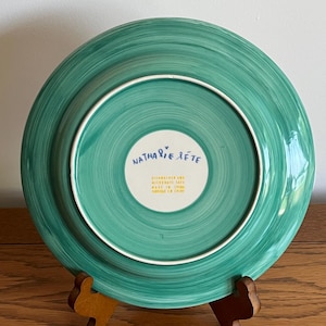 Może przedstawiać: Turkusowy ceramiczny talerz z białym obramowaniem i spiralnym wzorem. Sp&oacute;d talerza zawiera napis "Nathalie Lete", "Dishwasher and Microwave Safe" i "Made in China".