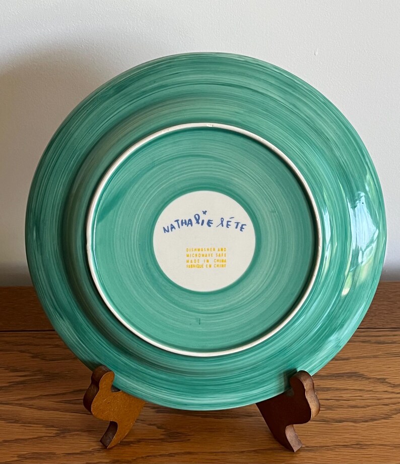 Może przedstawiać: Turkusowy ceramiczny talerz z białym obramowaniem i spiralnym wzorem. Sp&oacute;d talerza zawiera napis "Nathalie Lete", "Dishwasher and Microwave Safe" i "Made in China".