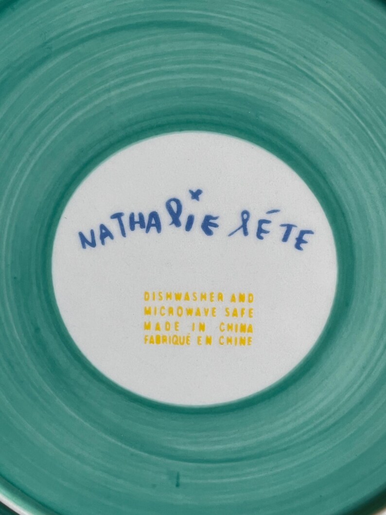 Może przedstawiać: Turkusowy i biały ceramiczny talerz z napisem "Nathalie Lete" w niebieskim skrypcie. Talerz ma r&oacute;wnież napis "Dishwasher and Microwave Safe Made in China" w kolorze ż&oacute;łtym.