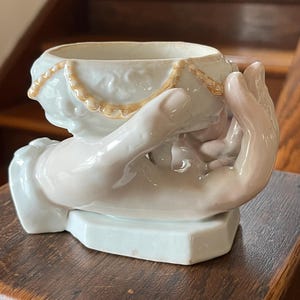 May include: A white ceramic planter in the shape of hands cradling a bowl. The bowl has a gold trim and a textured design. The hands are a pale pink color and the base is a light blue-green.