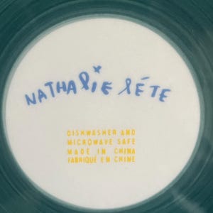 May include: A white ceramic plate with a teal green rim and a white center. The center features the name "Nathalie Lete" in blue script and text in yellow that reads "Dishwasher and Microwave Safe Made in China Fabriqu&eacute; en Chine."