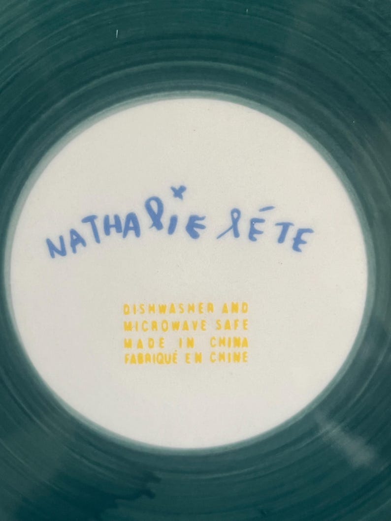 May include: A white ceramic plate with a teal green rim and a white center. The center features the name "Nathalie Lete" in blue script and text in yellow that reads "Dishwasher and Microwave Safe Made in China Fabriqu&eacute; en Chine."
