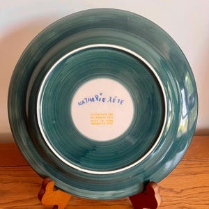 May include: A teal-colored ceramic plate with a white inner circle. The plate's underside features the text "Nathalie Lete" in blue script. The plate is displayed on a wooden stand. The plate is dishwasher and microwave safe.