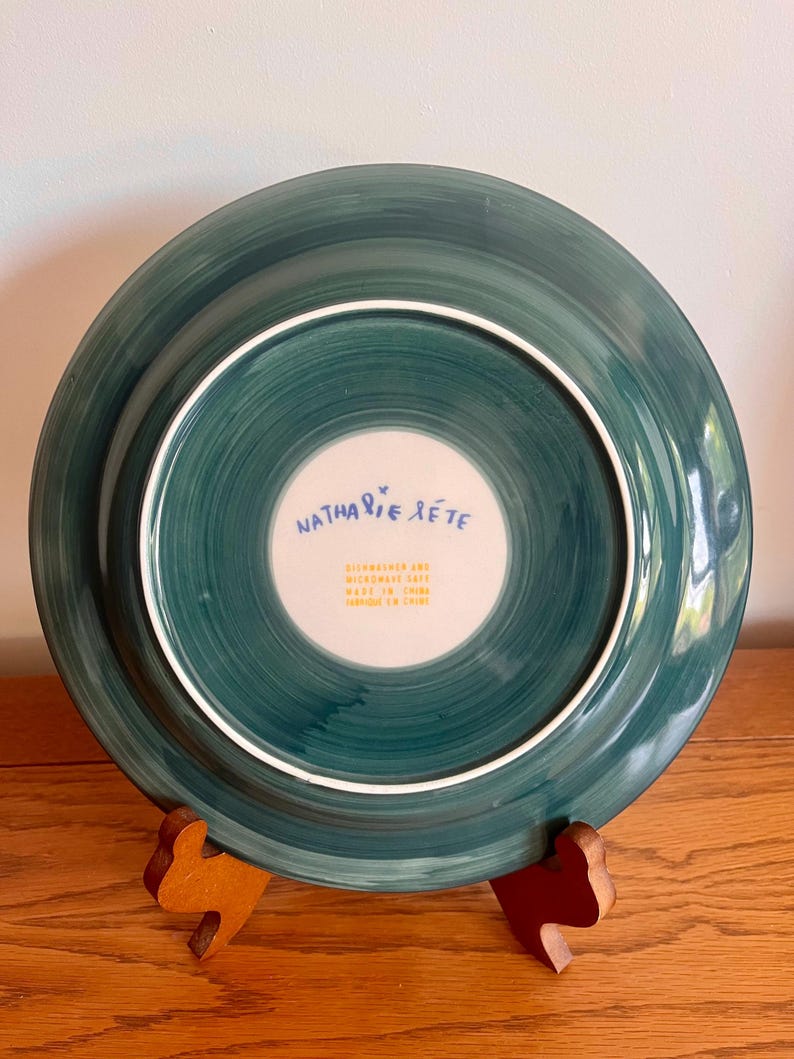 May include: A teal-colored ceramic plate with a white inner circle. The plate's underside features the text "Nathalie Lete" in blue script. The plate is displayed on a wooden stand. The plate is dishwasher and microwave safe.