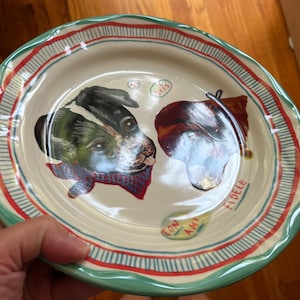Może przedstawiać: Kremowy ceramiczny talerz z ręcznie malowanym portretem psa. Talerz ma zieloną obw&oacute;dkę, czerwone i niebieskie paski oraz napisy "TON AMI" i "FIDELE". Pies ma czerwoną chustę.