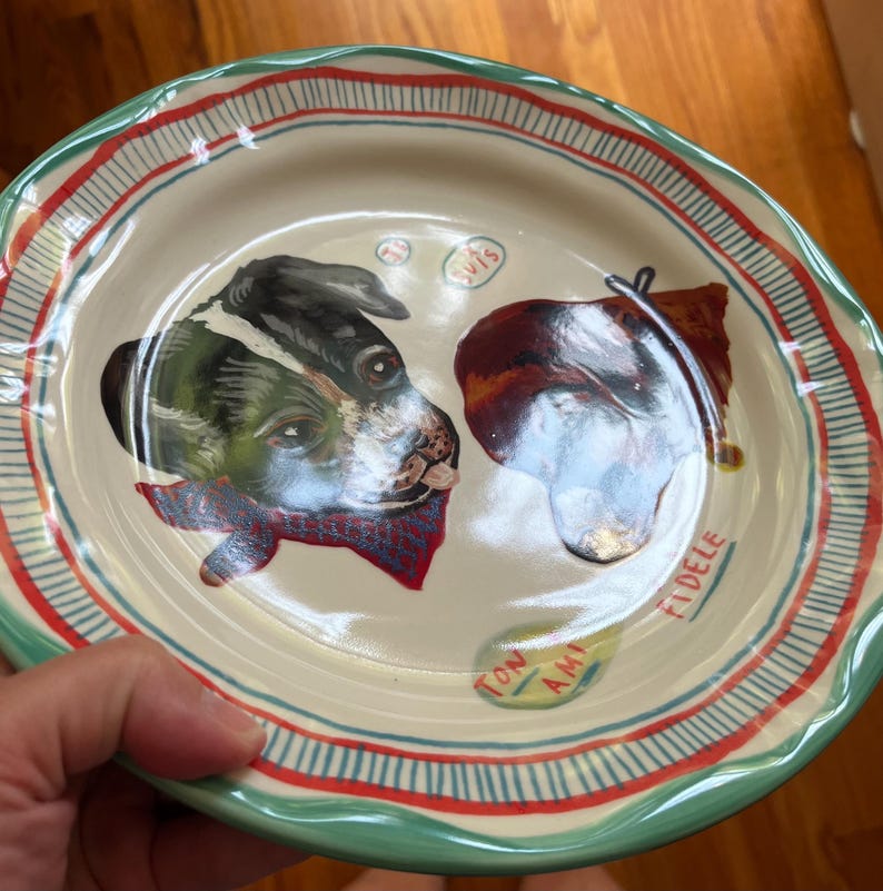 Może przedstawiać: Kremowy ceramiczny talerz z ręcznie malowanym portretem psa. Talerz ma zieloną obw&oacute;dkę, czerwone i niebieskie paski oraz napisy "TON AMI" i "FIDELE". Pies ma czerwoną chustę.