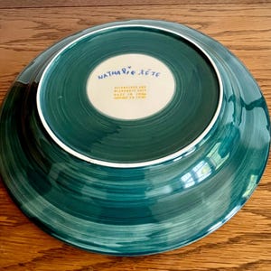 May include: A teal-colored ceramic plate with a white rim. The underside features the text "Nathalie Lete" in blue script and additional text in a smaller font. The plate has a spiral brushstroke design.